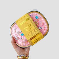 A hand with colorful beaded bracelets holds a $165 JRC Gift Bag from Jenna Rae Cakes against a plain light gray background.