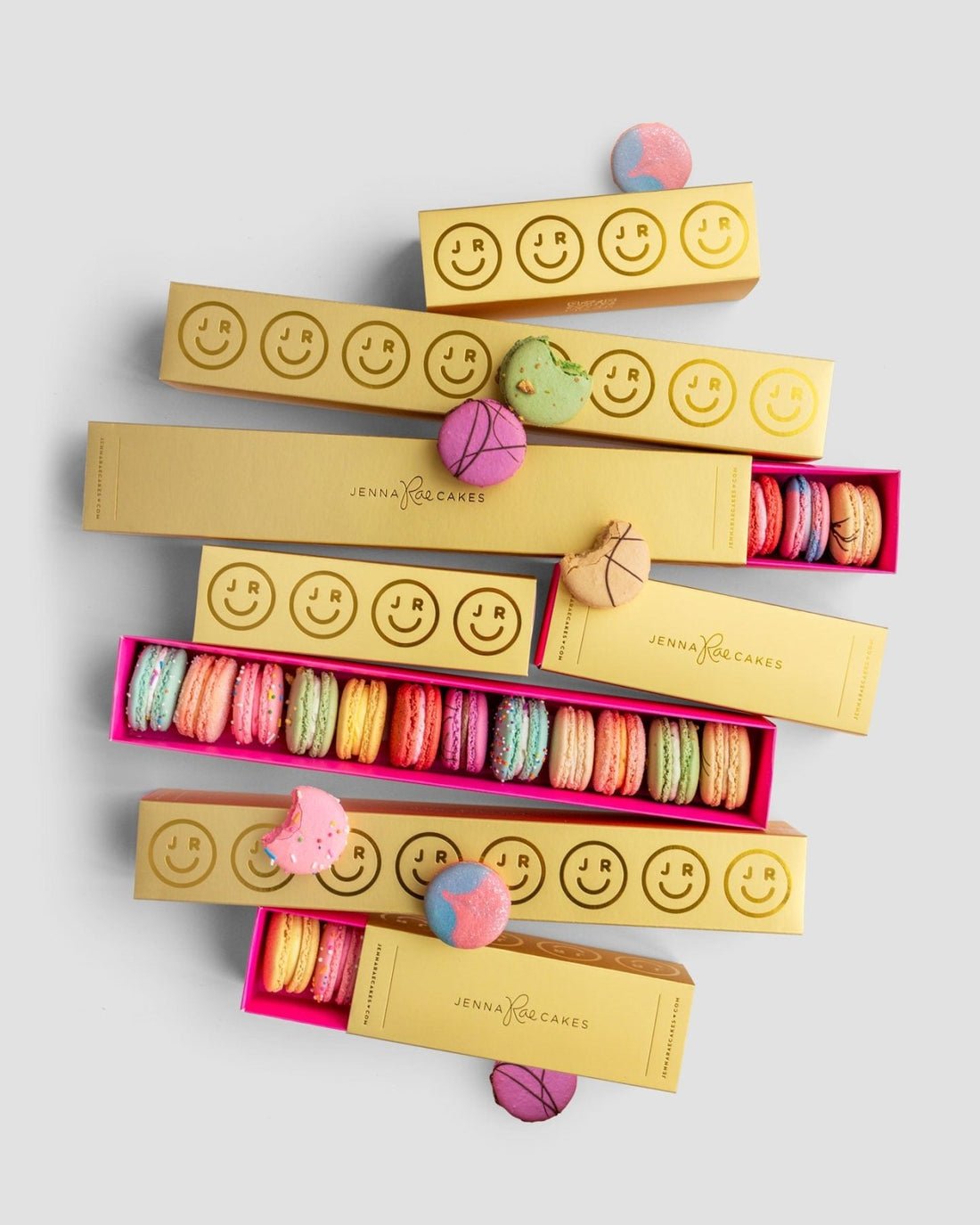 Jenna Rae Cakes $50 JRC Gift Bag features gold boxes with smiley faces, filled with colorful macarons and displayed on a light gray background, with extra loose macarons scattered around.
