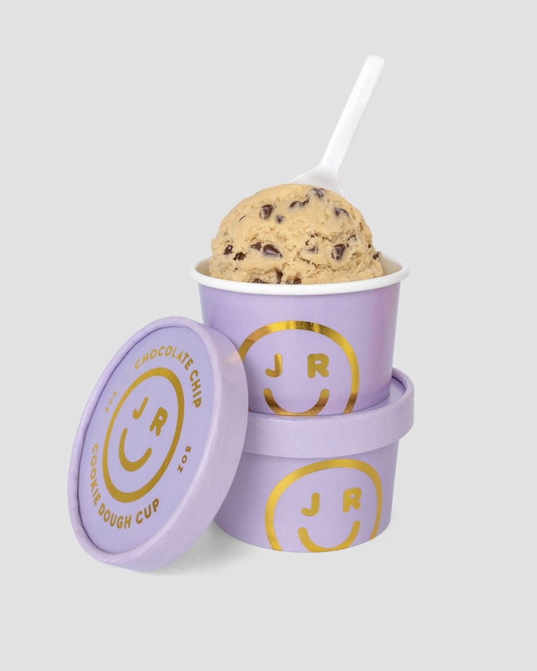 A $75 JRC Gift Bag by Jenna Rae Cakes sits atop a second purple cup, both with gold smiley faces and the JR logo. Nearby is a lid labeled Chocolate Chip Cookie Dough Cup. A white spoon rests in the cup.
