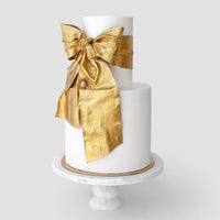 A two-tier white cake on a marble stand features a large bow on the top tier, shimmering with Jenna Rae Cakes Antique Gold Edible Lustre Dust (20g, pack of 3).