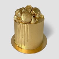 A cake adorned with Jenna Rae Cakes Antique Gold Edible Lustre Dust, shimmering gold icing, gold-dipped macarons, meringues, geometric toppers, and gold drip detail—elegantly set against a plain light background.