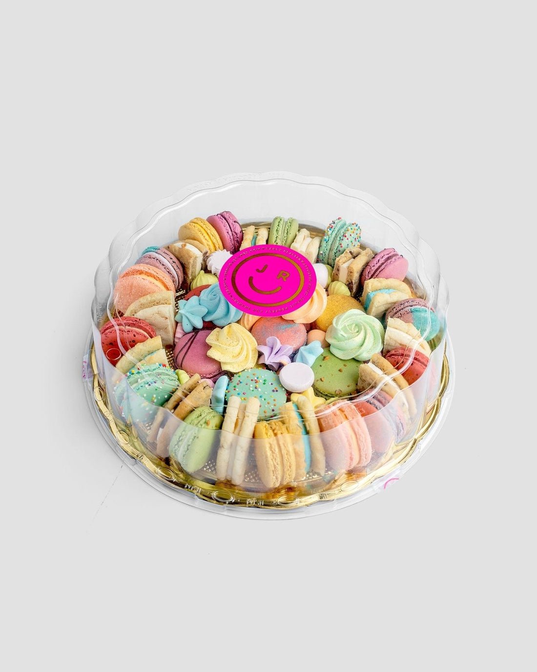 The Jenna Rae Cakes Assorted Party Platter features colorful macarons and meringue cookies in a clear round container, topped with a pink smiley-face sticker.