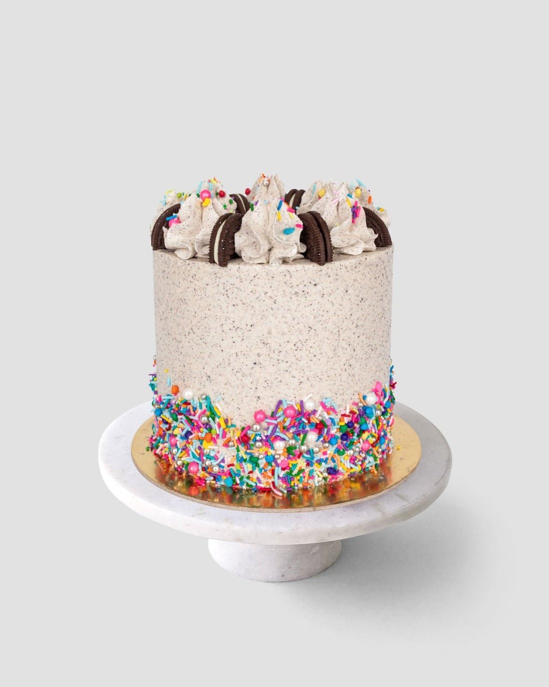 The Birthday Oreo Cake by Jenna Rae Cakes features cookies and cream frosting, whipped cream swirls, chocolate sandwich cookies on top, and colorful sprinkles around the base, beautifully displayed on a marble cake stand.