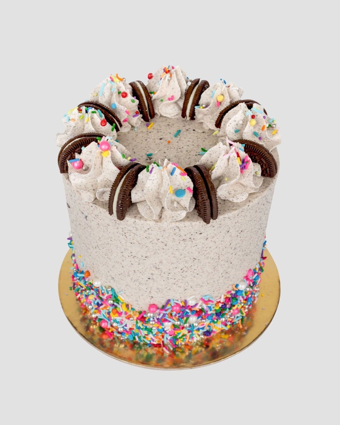 Shop Jenna Rae Cakes Birthday Oreo Cakes 101 In-Person Cake Decorating Class features a tall cookies and cream cake with swirled frosting, chocolate sandwich cookies, colorful sprinkles, and a sprinkle-covered base on a gold board.