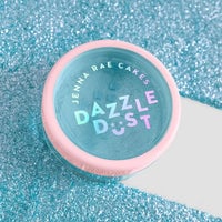A 5g shaker of Jenna Rae Cakes Blue Sky Edible Glitter, part of a 6-pack, with a pink lid and holographic lettering, sits on blue glitter.