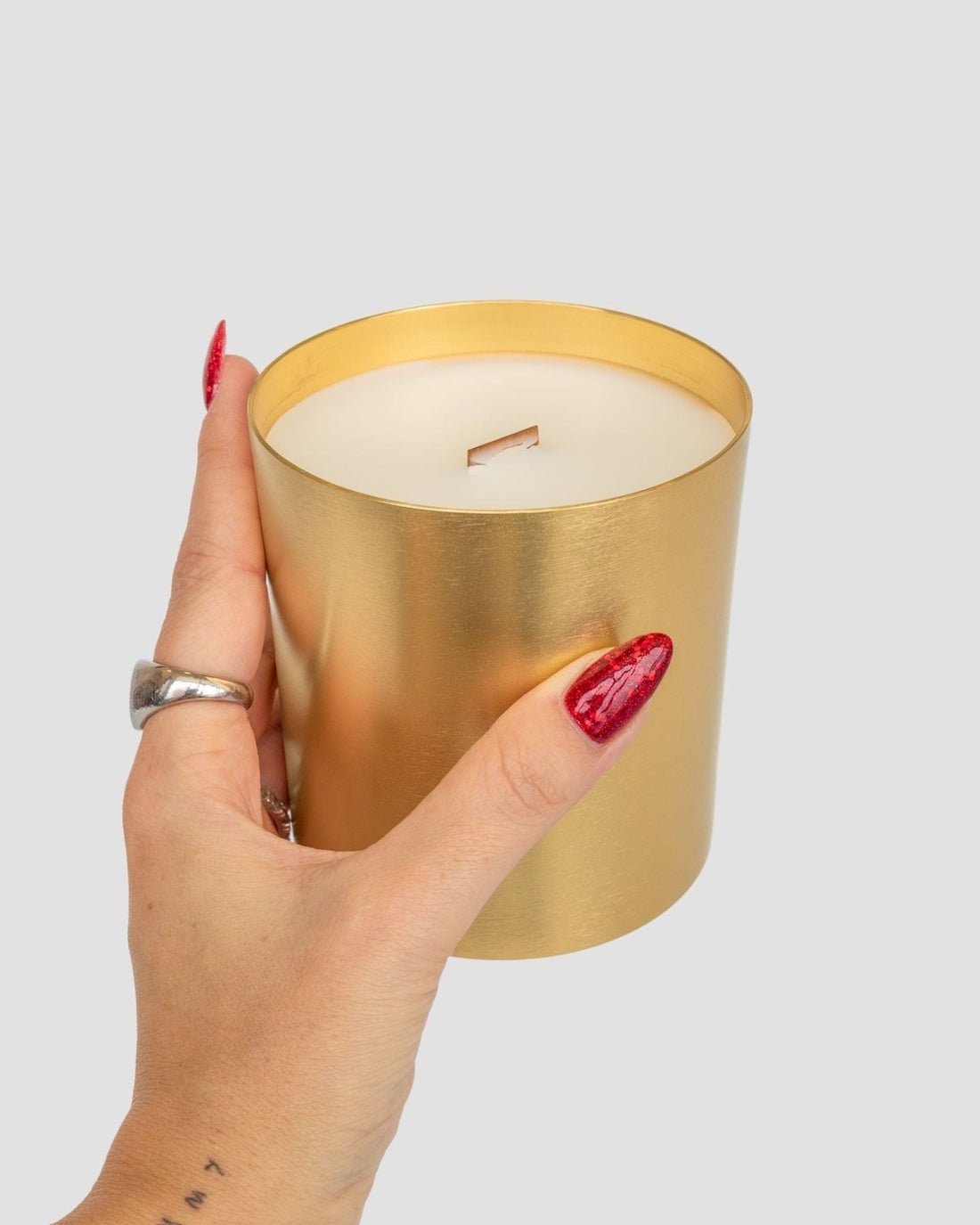A hand with red glittery nails and a silver ring holds the Jenna Rae Cakes Brown Butter Icing Candle, featuring a metallic gold finish and wooden wick, against a plain gray background.