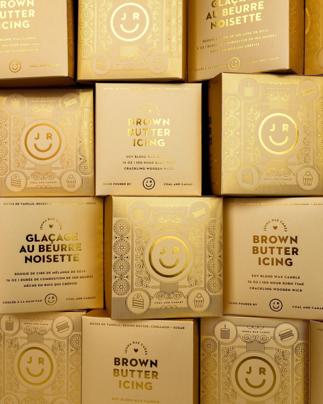 Several stacked golden boxes labeled Brown Butter Icing Candle from Jenna Rae Cakes, featuring a shiny finish, decorative patterns, a smiley face, and the JR logo.