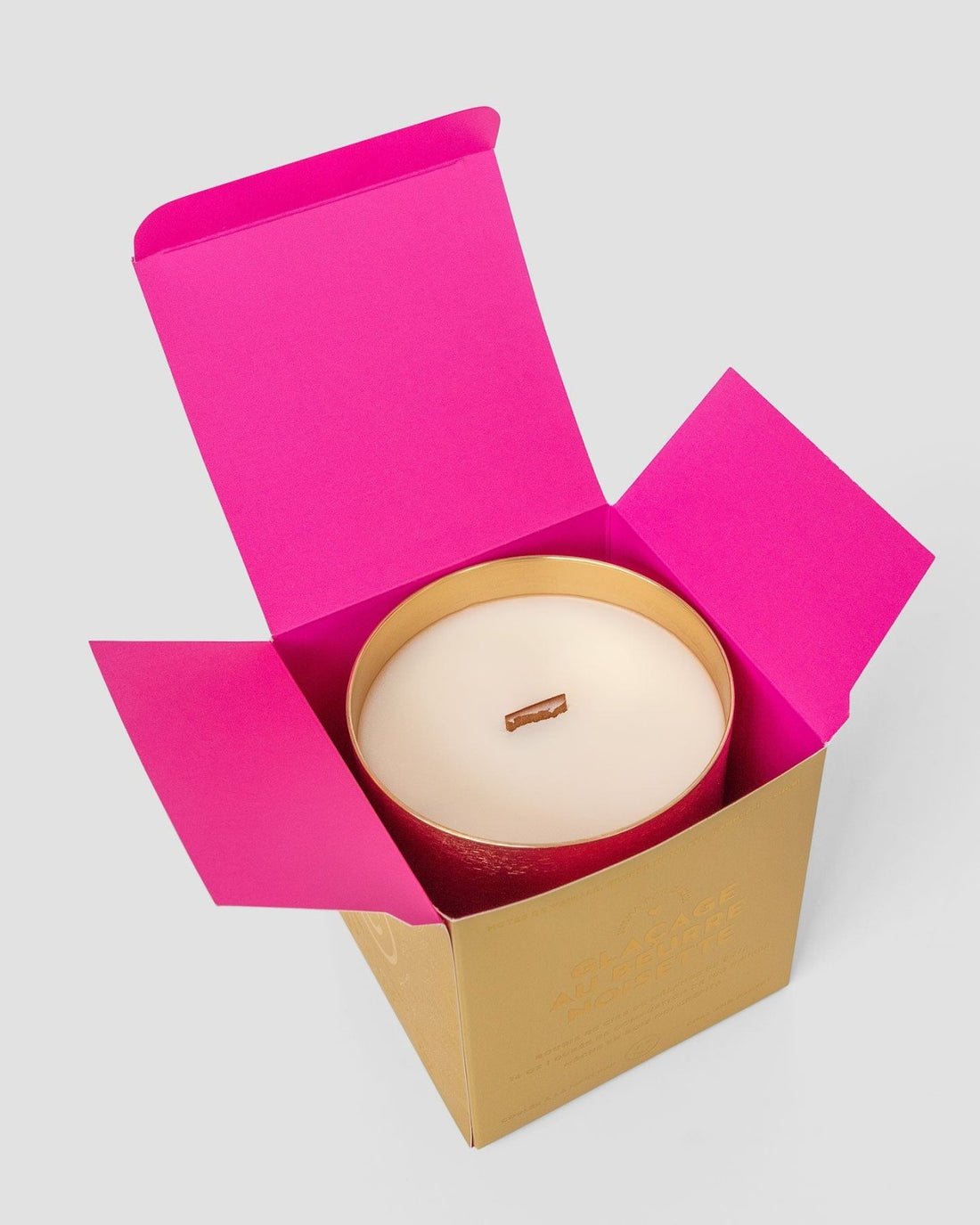 The Jenna Rae Cakes Brown Butter Icing Candle, a large white candle with a wooden wick in a gold container, is displayed in an open gold box with bright pink flaps on a light gray background.