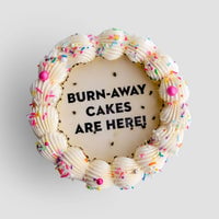A round Jenna Rae Cakes Burn Away Cake with white frosting, colorful sprinkles, and pink accents. The center displays BURN-AWAY CAKES ARE HERE! in black icing on a smooth white background.