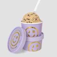 A scoop of Cookie Dough Cups by Jenna Rae Cakes in a purple cup with gold smiley face and JR branding, stacked on another matching cup, with a white spoon and matching lid nearby.