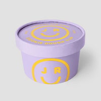 A lavender Jenna Rae Cakes Cookie Dough Cup with a matching lid, both featuring COOKIE DOUGH CUP in yellow and a smiley face with J R inside on the side and top.
