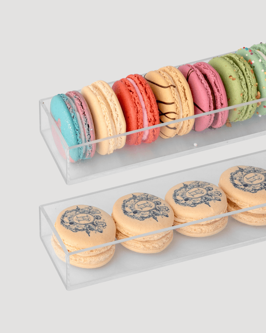 Two clear trays from Jenna Rae Cakes’ Dessert Tables feature colorful macarons in assorted flavors on top and beige macarons with intricate printed designs below, set against a plain light gray background.