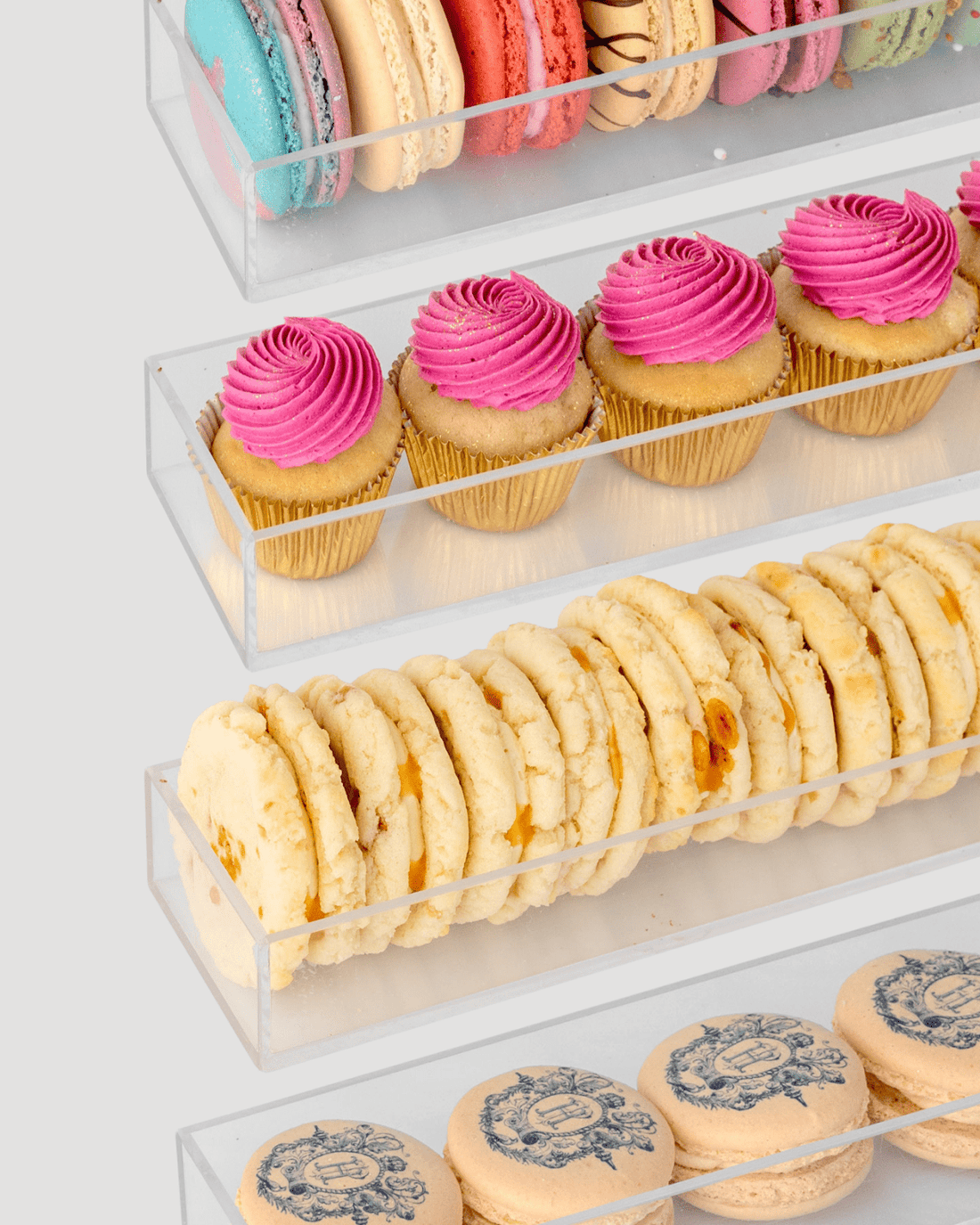 Jenna Rae Cakes Dessert Tables feature clear acrylic display shelves stocked with colorful macarons, bright pink cupcakes, crumbly cookies, and beige macarons with blue prints on a simple light background.