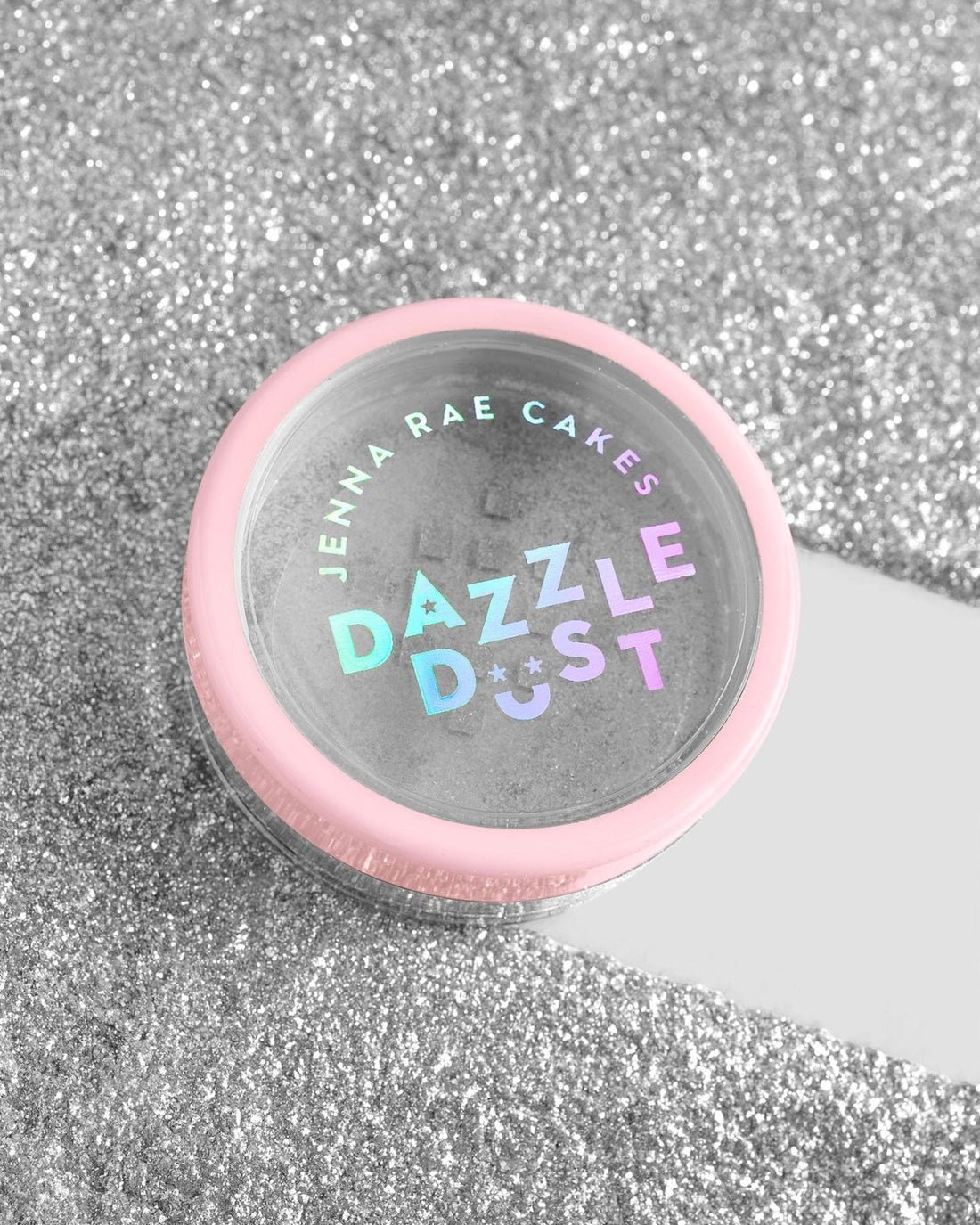 A small round container with a pink lid labeled “Jenna Rae Cakes Disco Ball Edible Glitter - 5g Shaker - Package of 6” sits on a sparkling silver glitter surface.