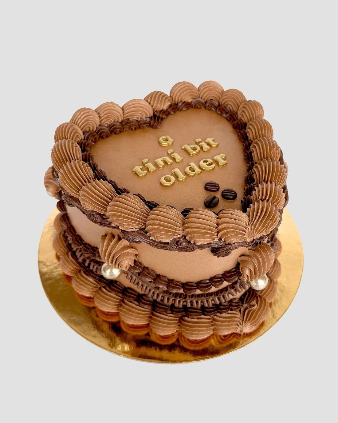 The Espresso Martini Heart Cake by Jenna Rae Cakes features piped chocolate frosting, gold pearls, and a tini bit older written in gold atop a heart-shaped cake on a gold cake board, set against a light background.