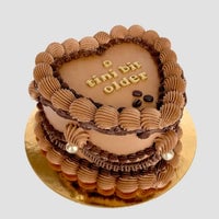 The Espresso Martini Heart Cake by Jenna Rae Cakes features piped chocolate frosting, gold pearls, and a tini bit older written in gold atop a heart-shaped cake on a gold cake board, set against a light background.
