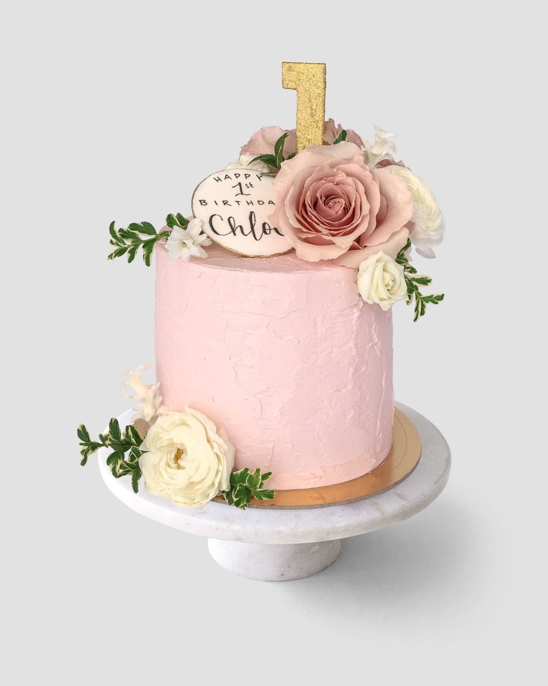 The Fresh Floral Buttercream Cake by Jenna Rae Cakes features pink frosting, fresh flowers, a gold glitter number 1 topper, and a round Happy 1st Birthday Chloe plaque, beautifully presented on a white cake stand.