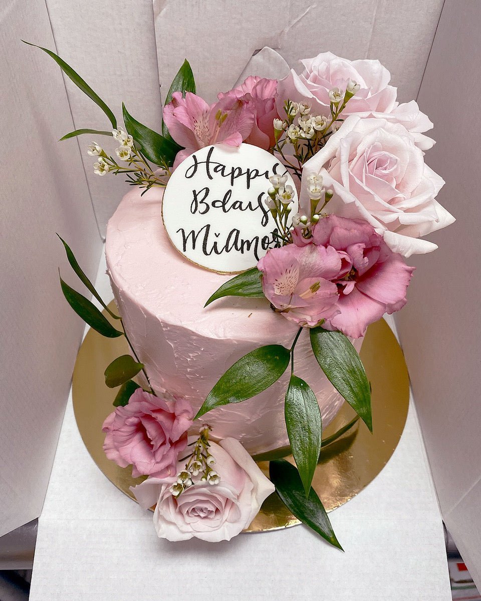 The Fresh Floral Buttercream Cake by Jenna Rae Cakes features pink frosting, fresh pink roses, green leaves, and a white Happy Bday Milana topper in black script, displayed on a gold cake board.