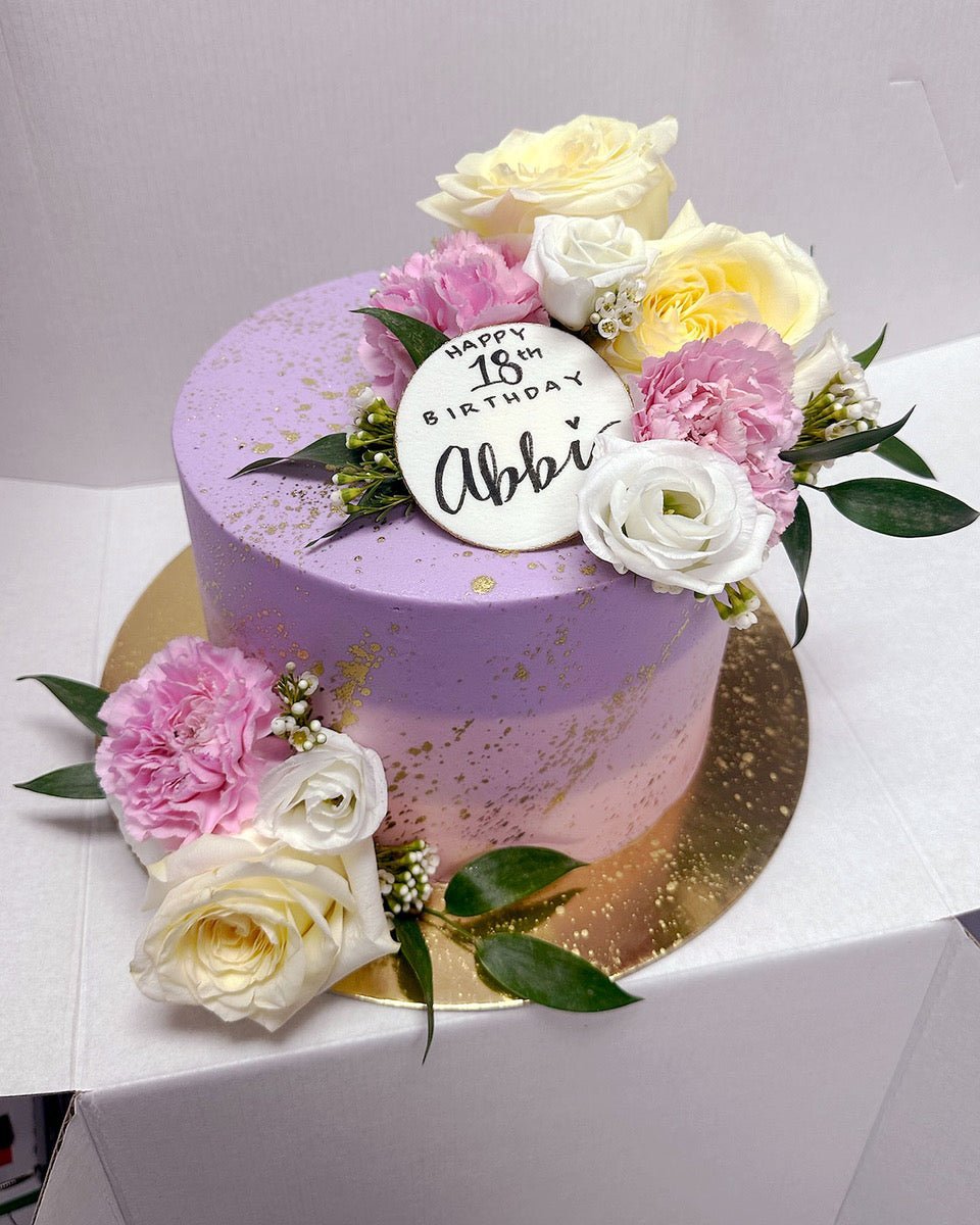 The Fresh Floral Buttercream Cake by Jenna Rae Cakes features a round purple design with gold splatters, pink and white flowers, green leaves, and a white disc on top reading Happy 18th Birthday Abbi in black. Presented on a gold board.