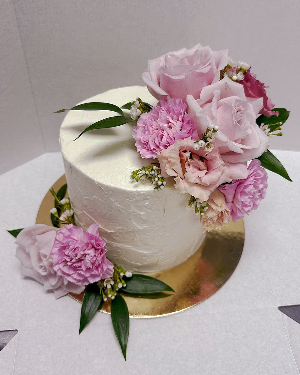The Fresh Floral Buttercream Cake by Jenna Rae Cakes features white frosting, pink roses, carnations, small white flowers, and green leaves on a gold cake board.