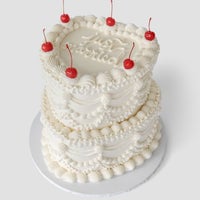 A two-tiered white cake decorated with ornate piped frosting, topped with six red cherries and the words Not Patriotic written on the top layer, set on a round white cake board against a plain background.