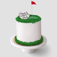 The Golf Ball Cake by Jenna Rae Cakes is a white frosted cake with green icing grass, topped with a red flag and a Happy Fathers Day plaque, displayed on a white cake stand.