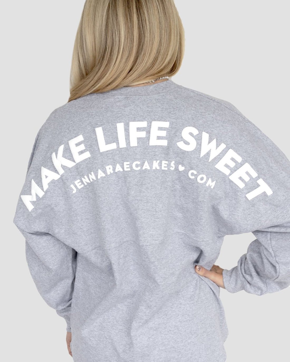 A person with long blonde hair wears the Grey + White Spirit Jersey by Jenna Rae Cakes, featuring white MAKE LIFE SWEET and JENNARAECAKES•COM text on the back, with one hand on their hip.