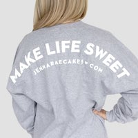 A person with long blonde hair wears the Grey + White Spirit Jersey by Jenna Rae Cakes, featuring white MAKE LIFE SWEET and JENNARAECAKES•COM text on the back, with one hand on their hip.