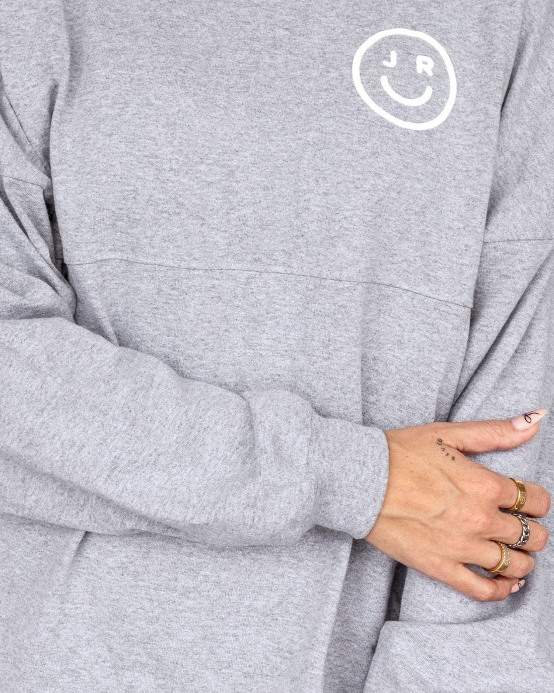 A person wears the Grey + White Spirit Jersey by Jenna Rae Cakes, featuring a white smiley face and the letters J and R on the chest, with their arms crossed and rings visible on their fingers.