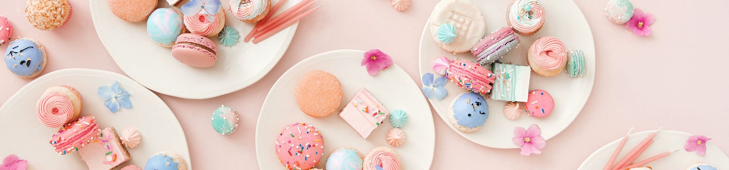 Five white plates on a pink surface, each arranged with pastel-colored treats like macarons, meringues, cookies, and candies. Pink and purple flower decorations are scattered around the plates.