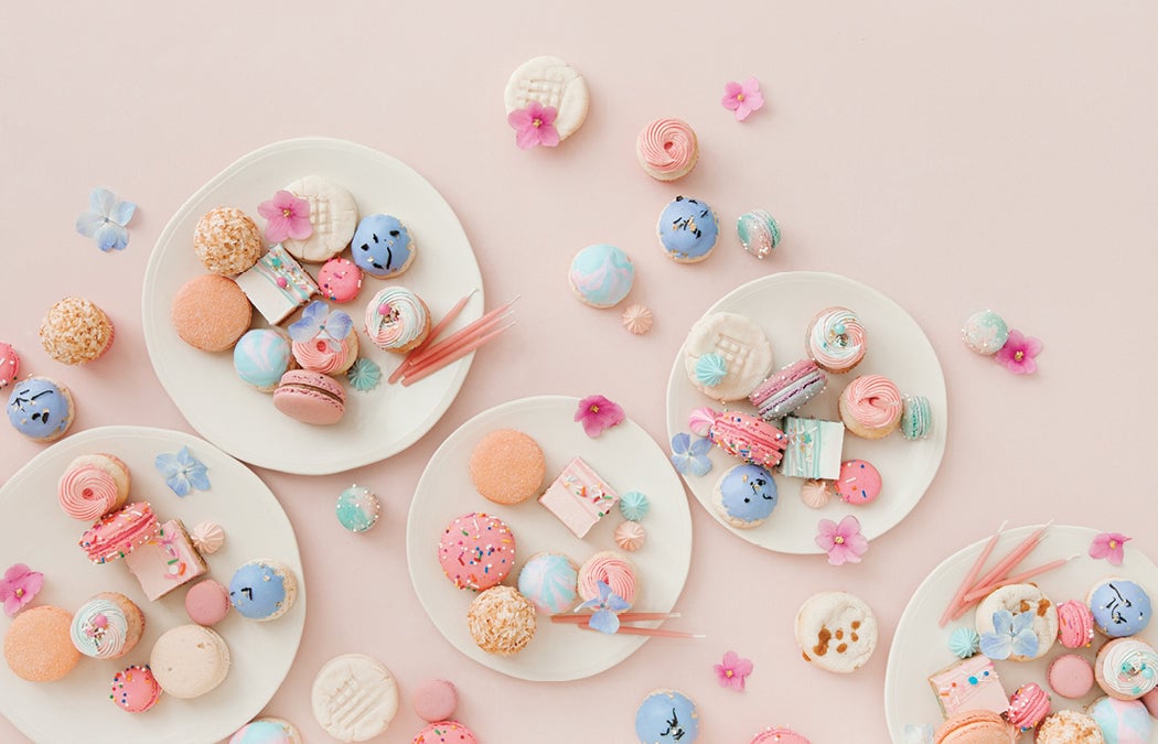 Five white plates filled with colorful pastel macarons, cookies, and sweets are arranged on a pink surface, surrounded by scattered confections and small decorative flowers in pink, purple, and blue tones.
