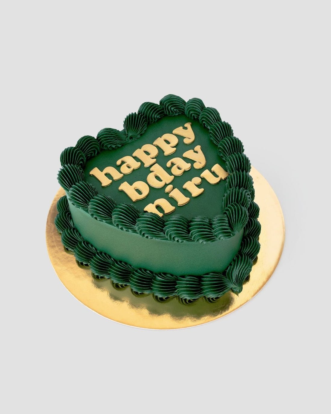 The Heart Cake by Jenna Rae Cakes features a dark green, heart-shaped design with wavy piped borders and “happy bday niru” in yellow icing, set on a gold board against a light background.