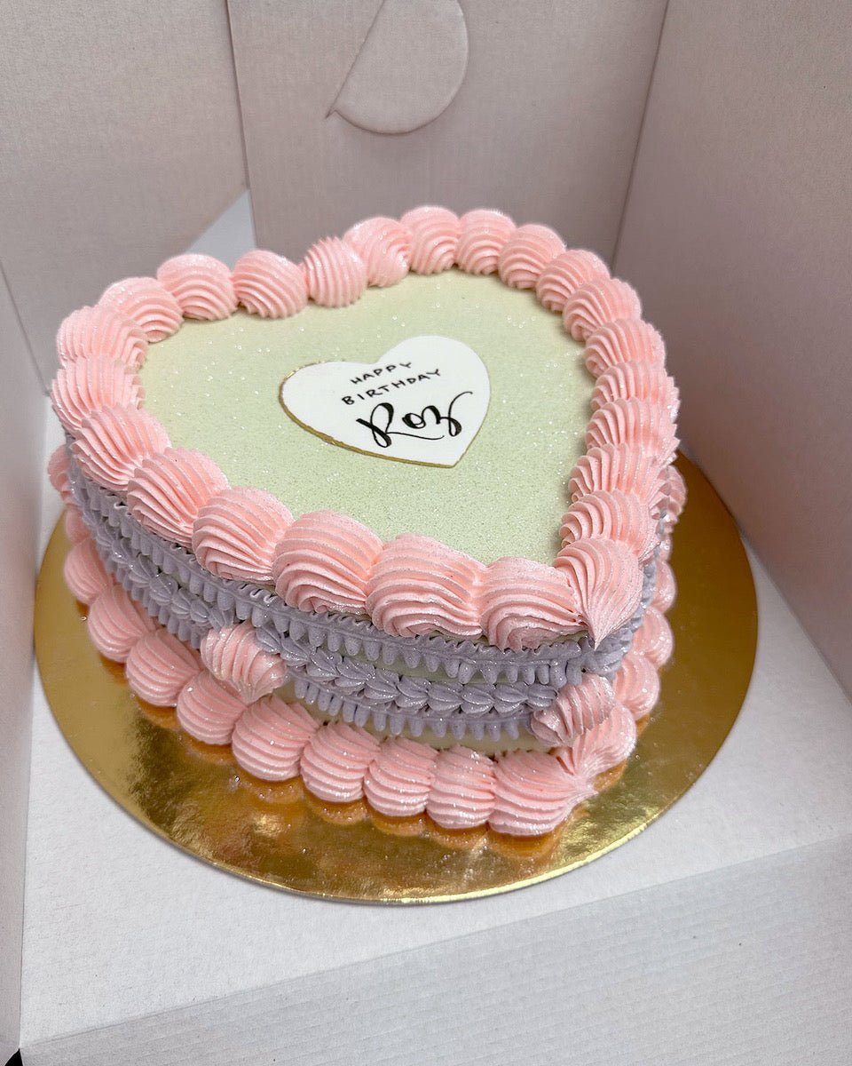 A heart-shaped cake with pink and purple icing borders on a gold base. A white heart plaque on top says Happy Birthday, Love in black script. The cake is inside an open white box.