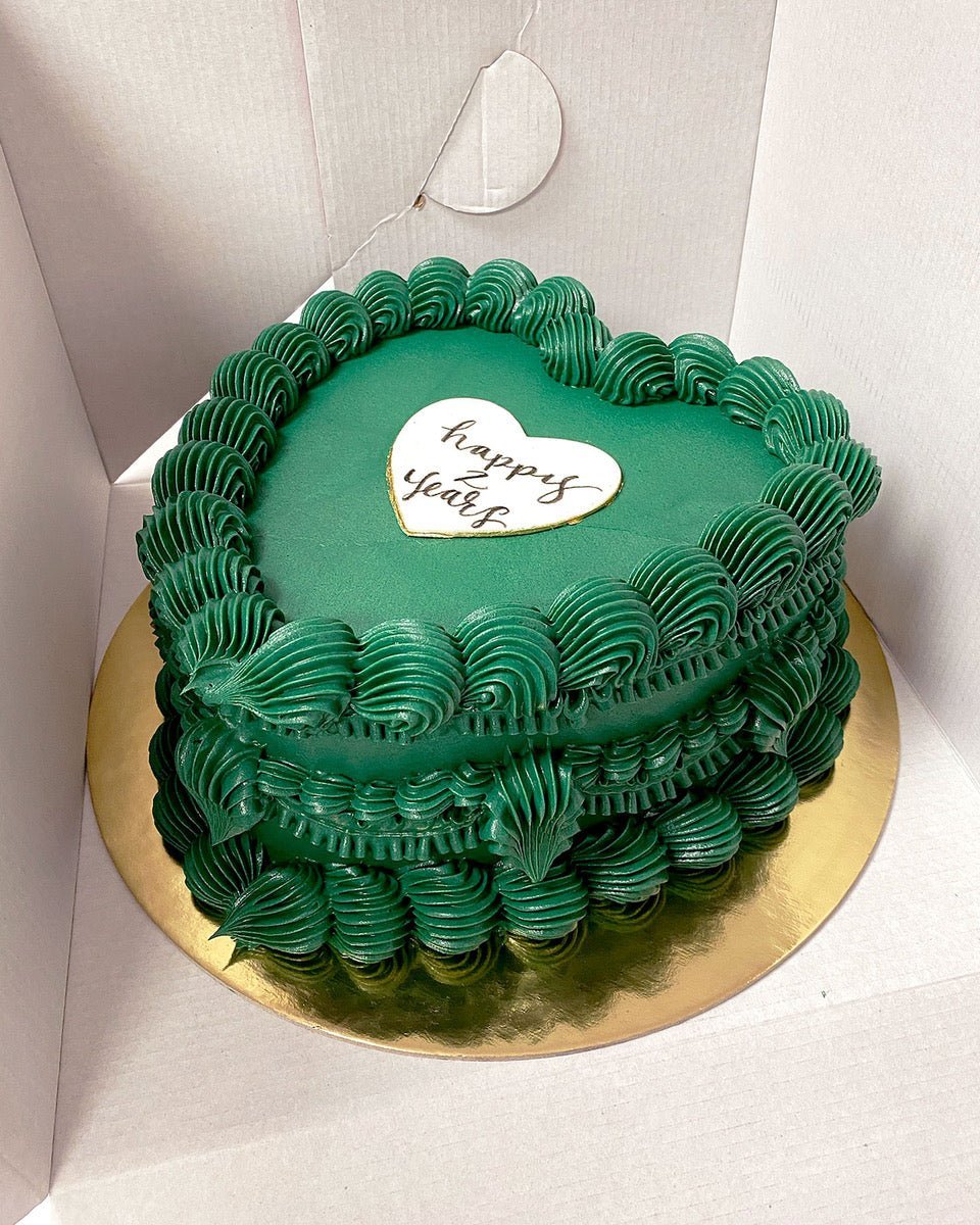 A heart-shaped cake with green frosting and decorative swirls, displayed on a gold cake board. A white heart-shaped plaque on top reads happy year in cursive, all inside a white cake box.