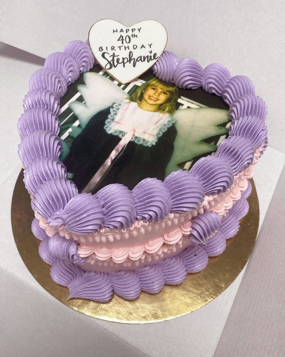 A heart-shaped cake with purple and pink frosting features a printed photo of a child in a fairy costume. A white heart topper on top reads Happy 40th Birthday Stephanie. The cake sits on a gold base.