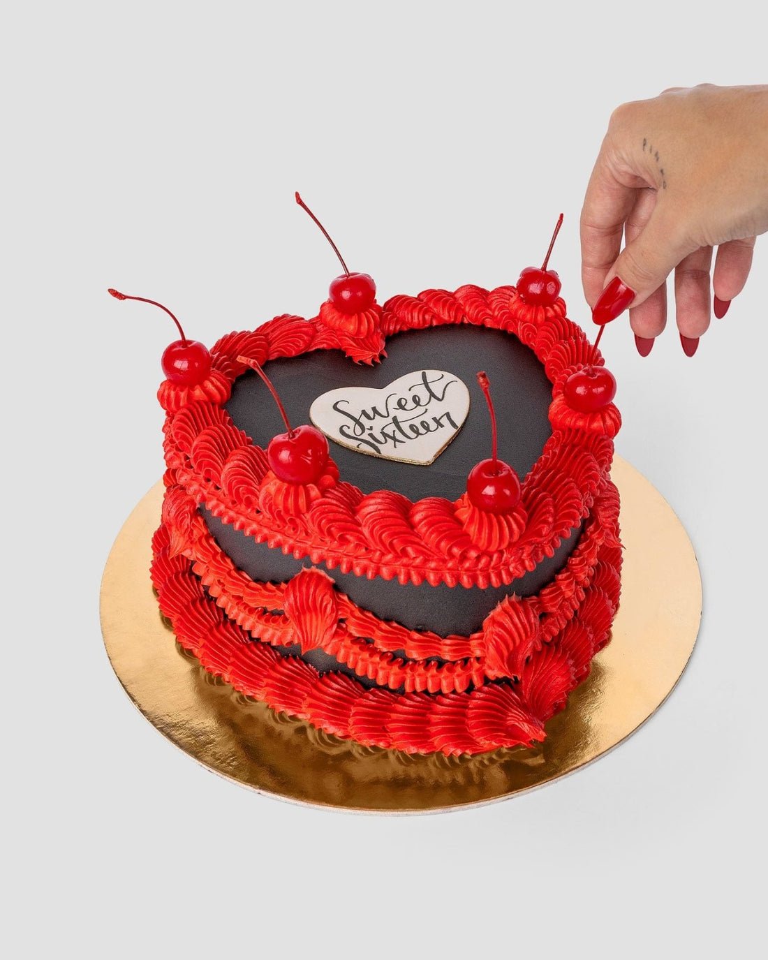 The Heart Cake by Jenna Rae Cakes features red frosting, a black icing center with Sweet Sixteen, topped with cherries. A hand places a cherry on top as the cake sits on a gold board against a light background.