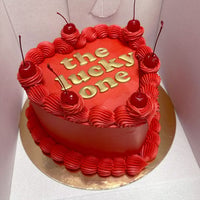 A heart-shaped red cake with red frosting, topped with five cherries and the words the lucky one written in gold letters, placed on a gold cake board inside a white box.