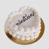 The Heart Cake by Jenna Rae Cakes is a white, heart-shaped cake with decorative icing borders and nineteen written in black icing, set on a round gold cake board.