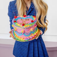 A woman in a blue houndstooth dress holds a pink, heart-shaped cake adorned with colorful icing swirls and cherries, crafted using the Jenna Rae Cakes x Colour Mill Pack by Jenna Rae Cakes. Her long blonde hair falls over her shoulders.