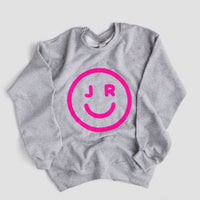 The JRC Smiley Sweatshirt by Jenna Rae Cakes is a gray top with a bold pink smiley face and the letters J and R as eyes printed on the front.