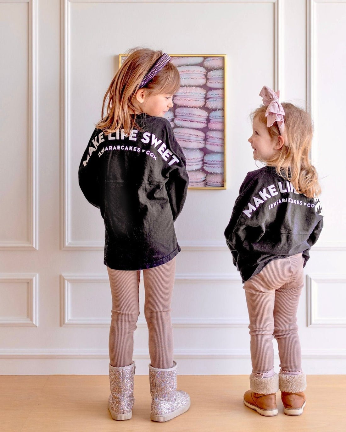 Two young girls smile at each other in front of a white wall and macaron photo, both wearing matching Jenna Rae Cakes Kids Black Spirit Jersey shirts, tan leggings, boots, and headbands.