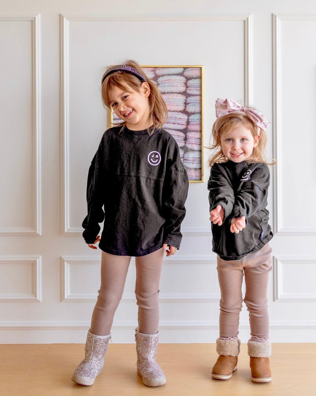 Two young girls indoors smile in matching Jenna Rae Cakes Kids Black Spirit Jerseys, tan leggings, and boots. One wears a headband; the other sports a large bow headband. Pastel artwork is displayed behind them.