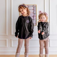 Two young girls indoors smile in matching Jenna Rae Cakes Kids Black Spirit Jerseys, tan leggings, and boots. One wears a headband; the other sports a large bow headband. Pastel artwork is displayed behind them.