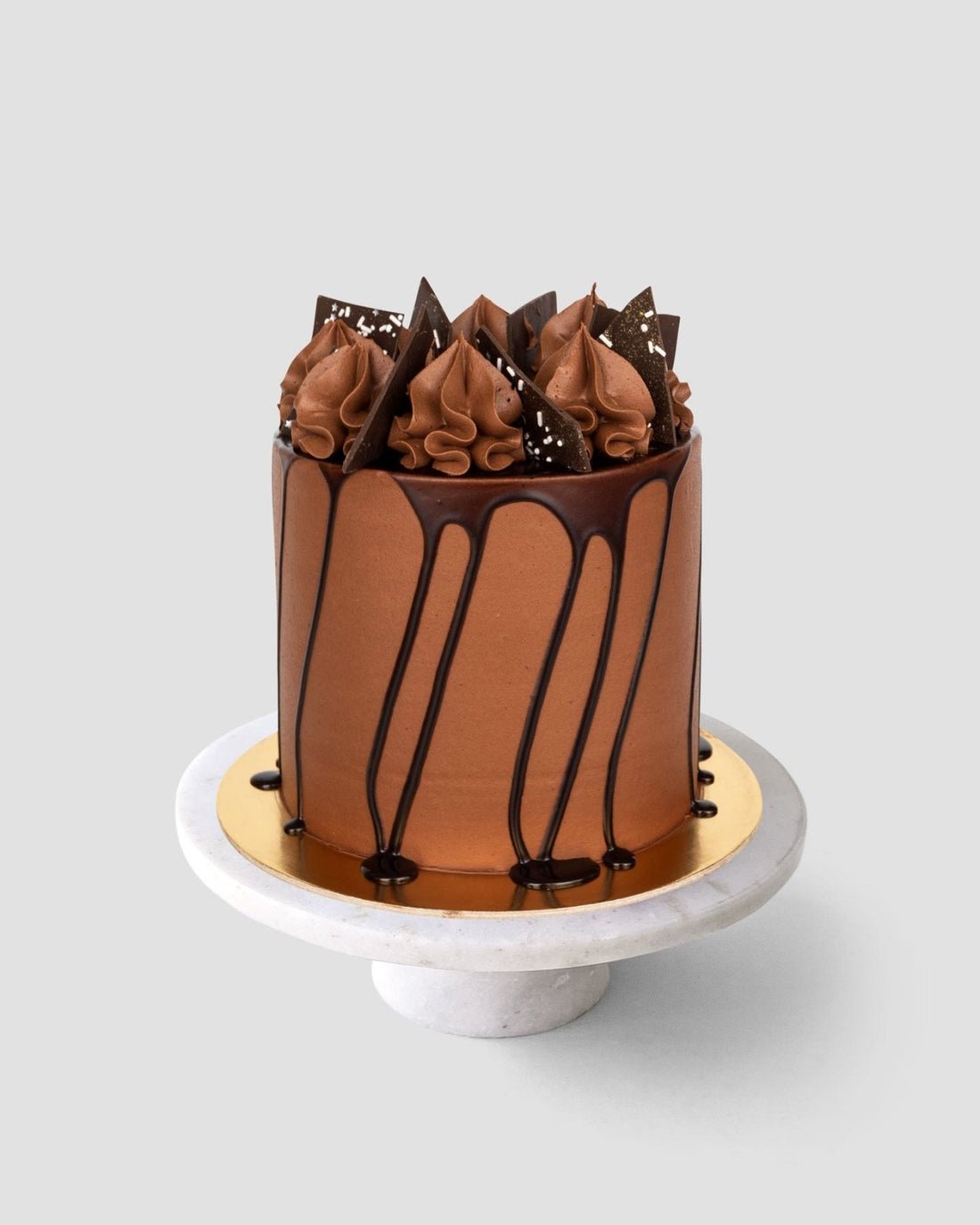 A tall chocolate cake from Jenna Rae Cakes’ Last Minute Cakes sits on a marble stand with a gold base, featuring smooth frosting, ganache drips, chocolate swirls, and triangles, against a plain gray background.