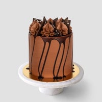 A tall chocolate cake from Jenna Rae Cakes’ Last Minute Cakes sits on a marble stand with a gold base, featuring smooth frosting, ganache drips, chocolate swirls, and triangles, against a plain gray background.