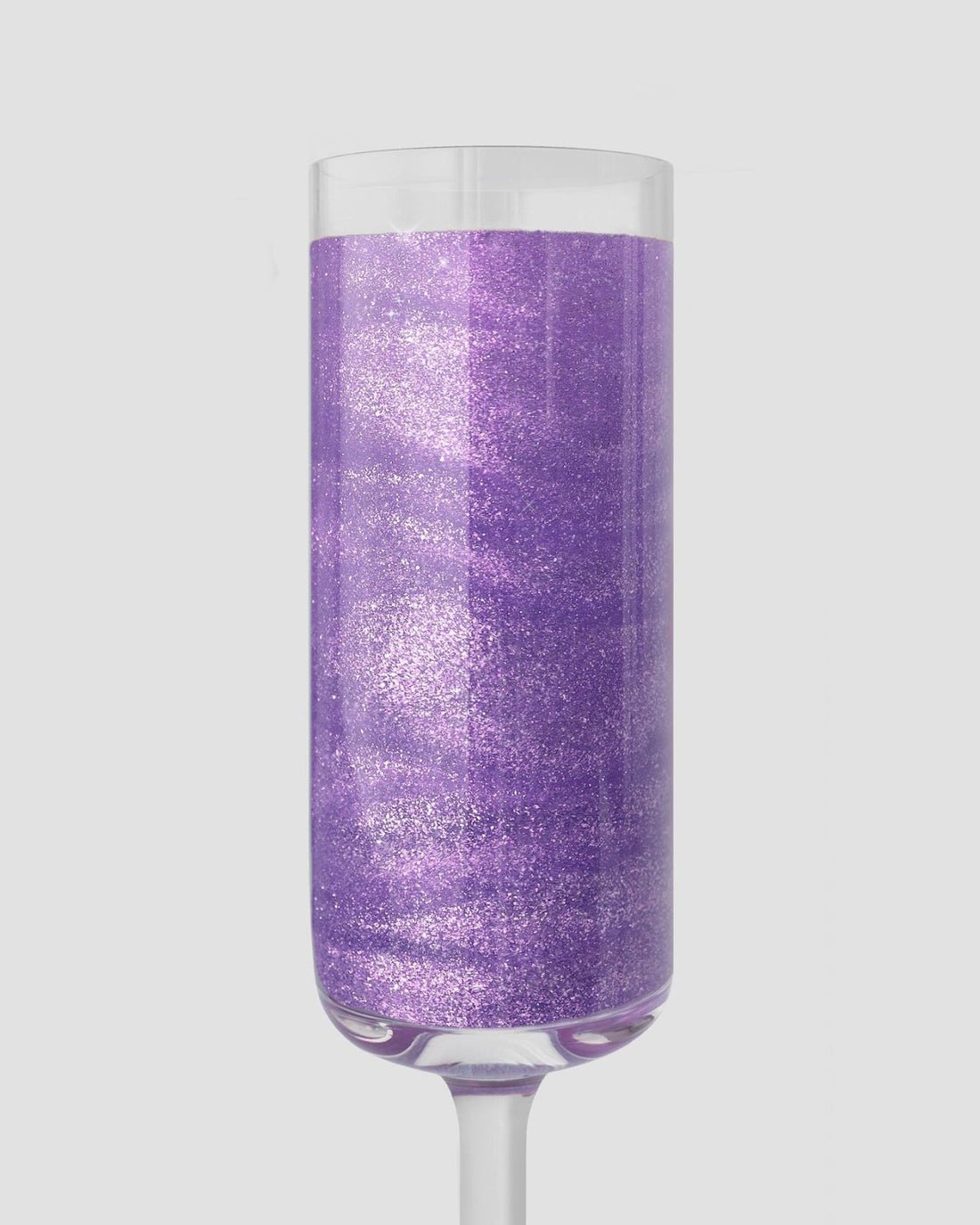 A clear champagne flute contains sparkling, shimmery purple liquid made with Jenna Rae Cakes Lavender Haze Edible Glitter (5g Shaker, Package of 6) against a plain light gray background.