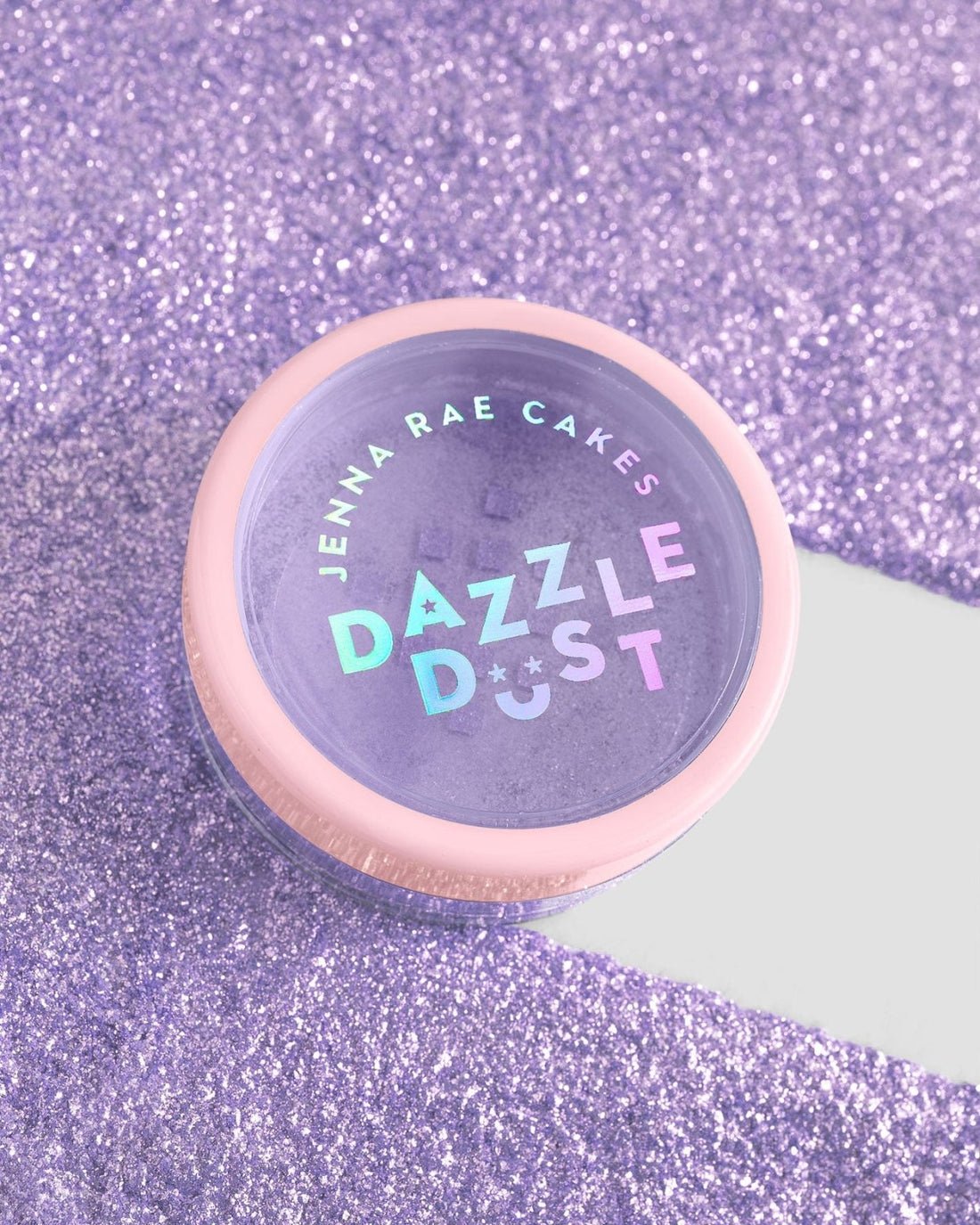 A package of six 5g shakers of Jenna Rae Cakes Lavender Haze Edible Glitter sits on a sparkling purple surface. Each shaker features a light pink lid and holographic lettering.