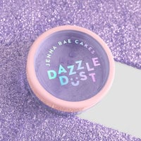 A package of six 5g shakers of Jenna Rae Cakes Lavender Haze Edible Glitter sits on a sparkling purple surface. Each shaker features a light pink lid and holographic lettering.