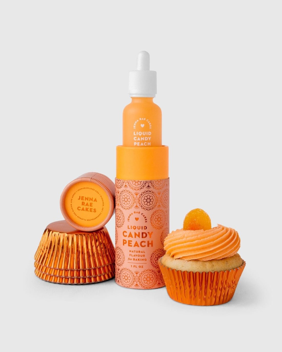 A bottle of Jenna Rae Cakes Liquid Candy Peach, featuring an orange label, is placed by its box, two stacked orange cupcake liners, and a peach-frosted cupcake with a small orange candy on a light backdrop.