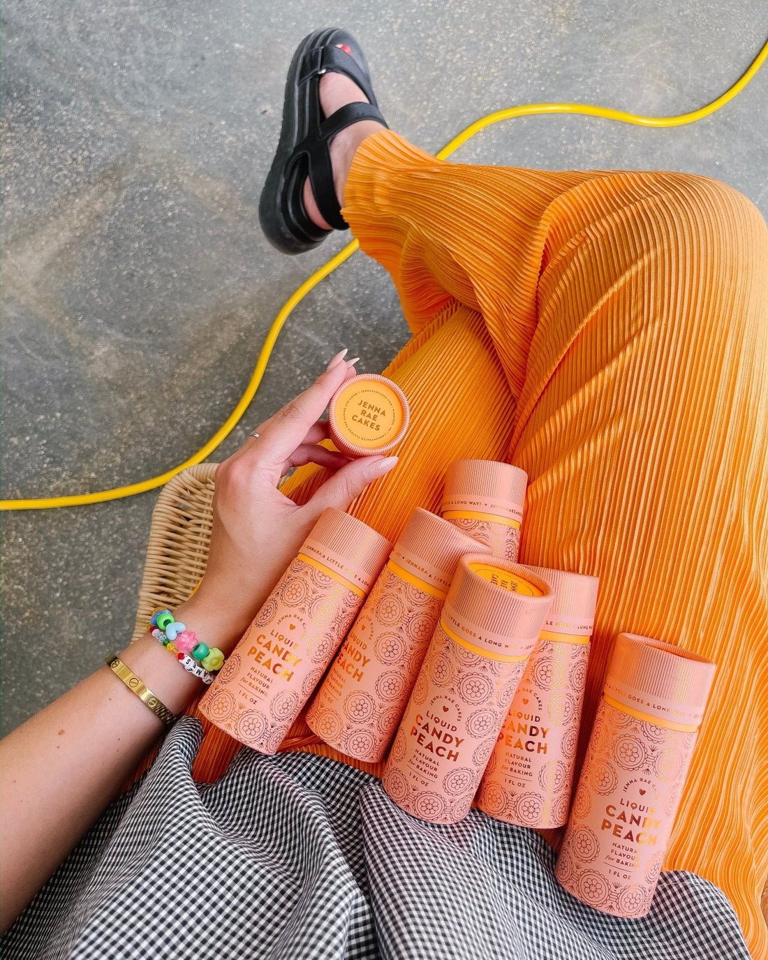 Wearing bright orange pleated pants, a person holds a round container of Jenna Rae Cakes Liquid Candy Peach and has several matching tubes on their lap, accessorized with bracelets and black sandals beside a yellow cord.